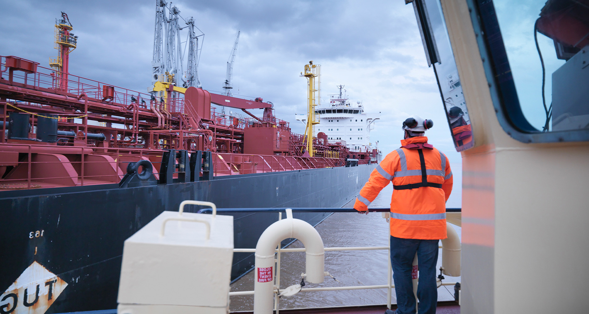 Worker in orange jacket observes large red industrial ship at port.