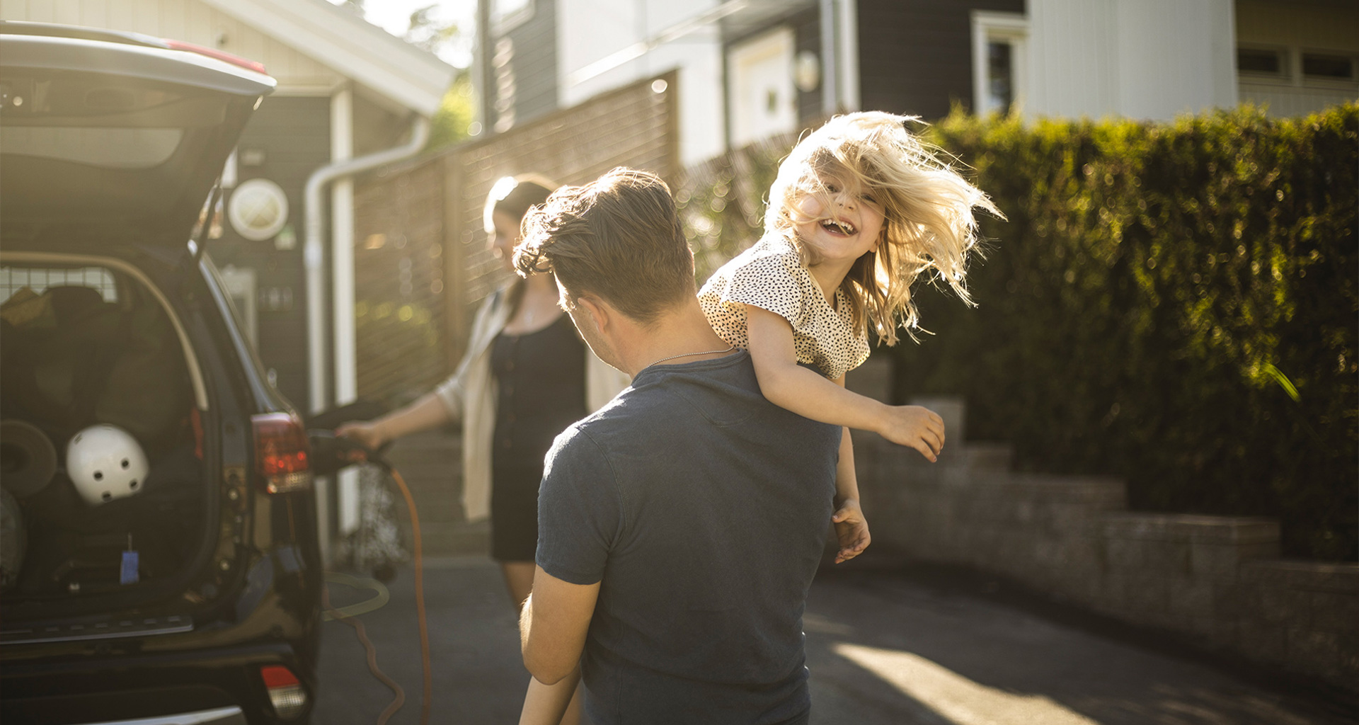 Man carrying smiling child near an open car trunk in a sunny driveway.