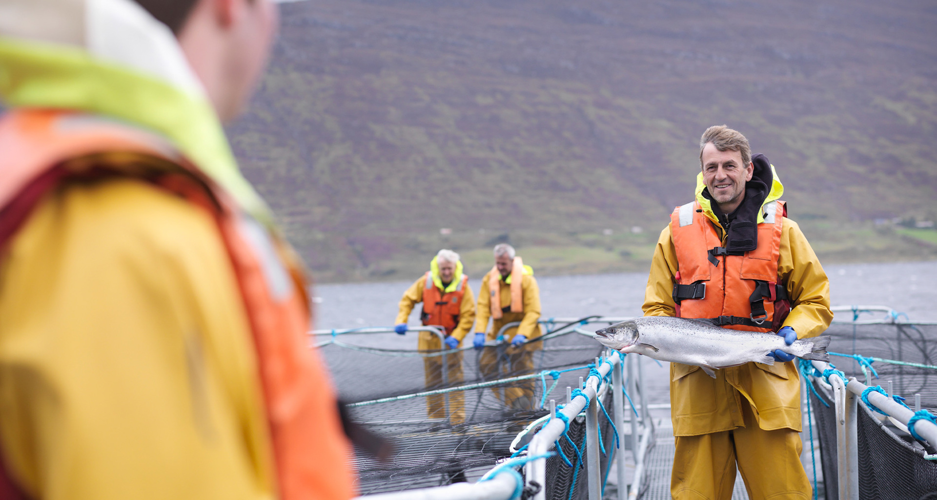 Man in orange life jacket holding large fish on aquaculture farm.